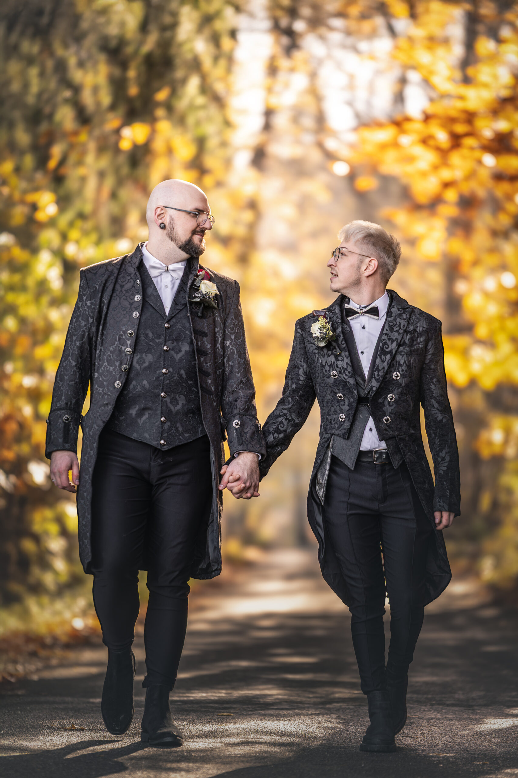 Homosexuelles Paar im viktorianischen Kleidungsstil geht Hand in Hand im Herbstwald auf die Linse von Kameralaus zu.