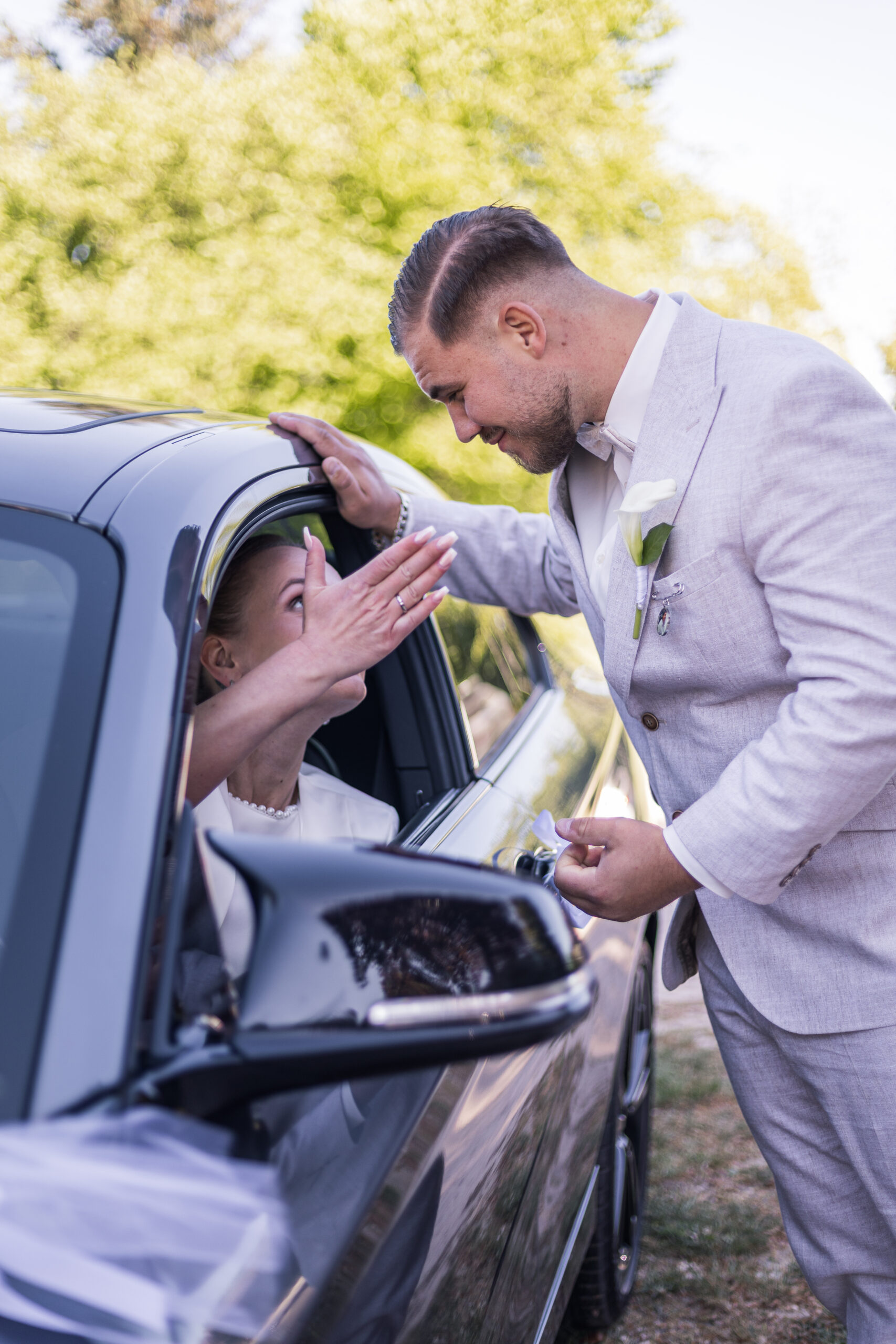 Frisch vermähltes Paar, Braut sitzt in ihrem BMW und droht ihrem frischen Ehemann amüsiert, fotografiert durch Kameralaus.